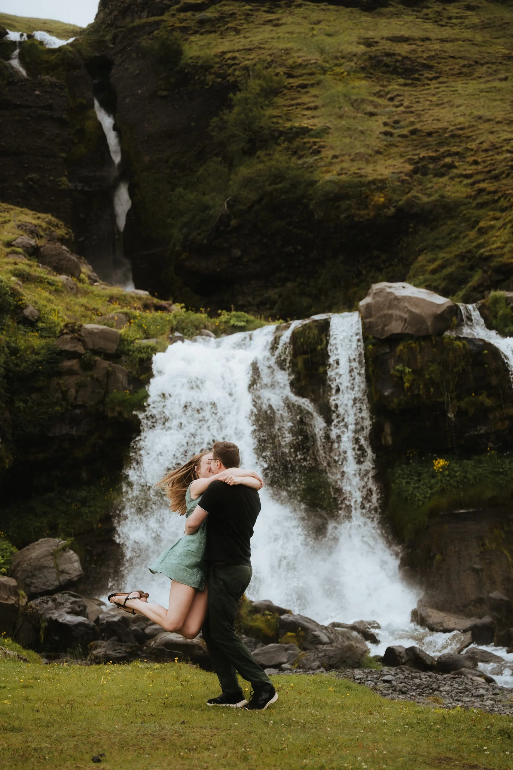 The couple embracing mid-spin in front of a cascading waterfall on a green mountainside