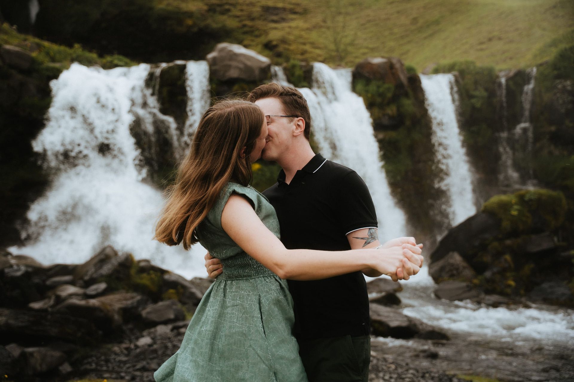 The couple kissing in front of a wide curtain of waterfalls on a green mountainside