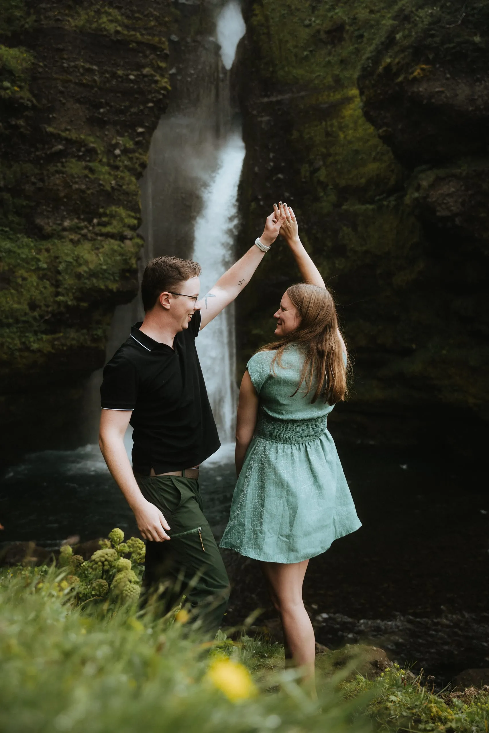 The couple laughing and dancing hand in hand before a waterfall in a mossy gorge