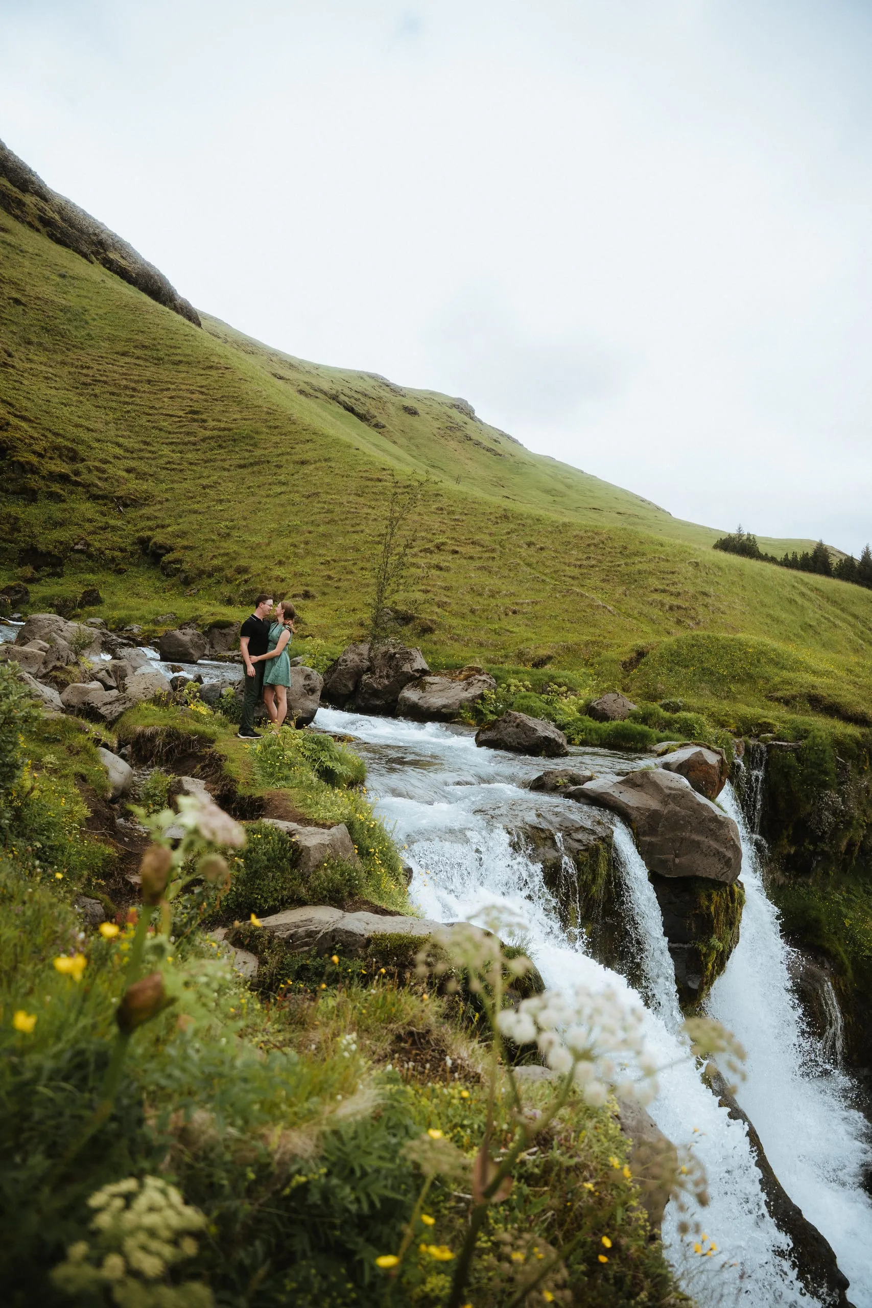 The couple standing beside a mountain stream and waterfall beneath a wide open sky