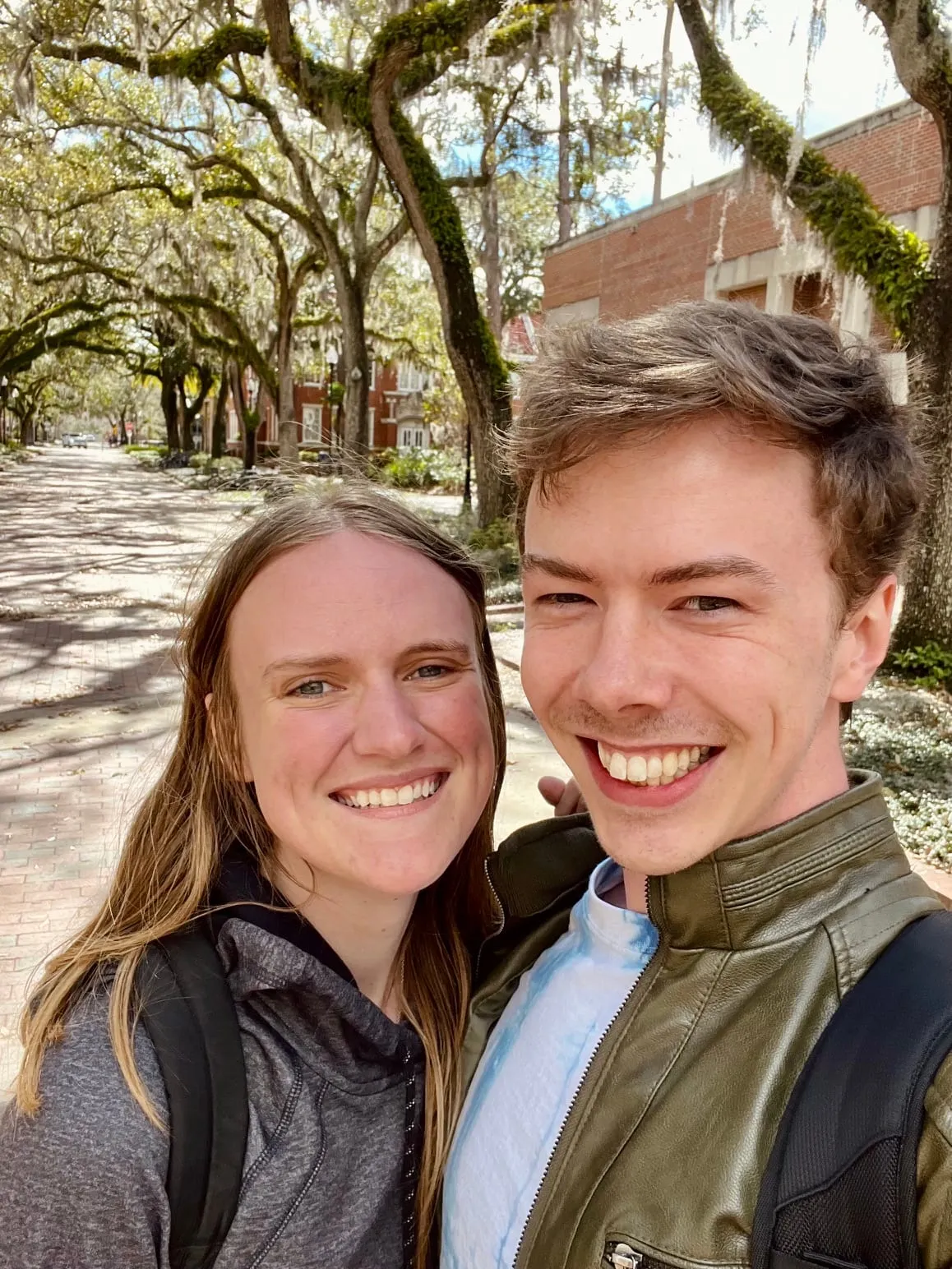 The couple taking a sunny selfie under moss-draped oak trees on a campus path